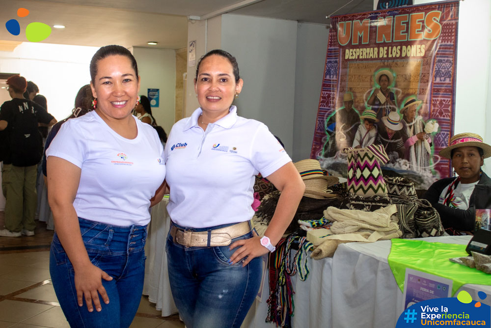 Coordinadora de la Unidad de Emprendimiento, Magaly Cerón y la docente Mabel Irene Paz, sonriendo al lado de un emprendimiento invitado a la feria.